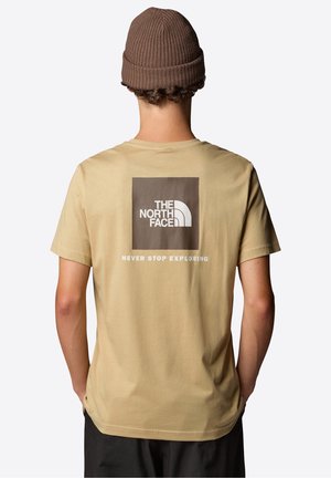 Osoba nosząca beżowy t-shirt The North Face z logo i tekstem "Never Stop Exploring" na plecach oraz brązową dzianą czapkę.