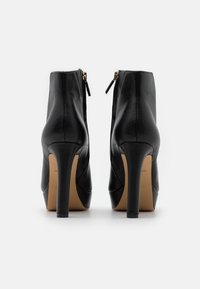 Botines de piel negra con tacón alto, textura suave, cierre de cremallera en la parte trasera y suela de color marrón claro. Forma elegante con diseño de punta afilada.