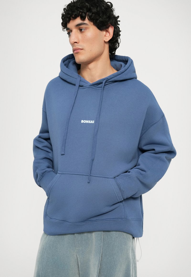 Hoodie en coton bleu avec poche kangourou, cordons de serrage et texte "BONSAI" en blanc, présentant une coupe décontractée et une texture douce.