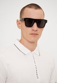 Sorte firkantede solbriller med mørke linser, med en gullakkent på templene. Båret sammen med en hvit polo skjorte med svart kant.