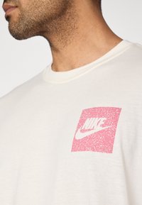 Μπεζ βαμβακερό t-shirt με λογότυπο Nike σε ροζ και λευκό, σχεδιασμένο σε τετράγωνο στο αριστερό μέρος του στήθους.