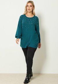 Pull long sans manches turquoise avec un col rond, orné de décorations métalliques éparpillées. Associé à un pantalon skinny noir et des bottines.