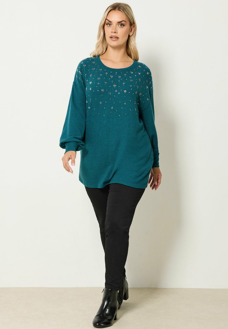 Pull long sans manches turquoise avec un col rond, orné de décorations métalliques éparpillées. Associé à un pantalon skinny noir et des bottines.