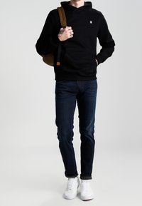 Sudadera negra con capucha y un bolsillo frontal, combinada con jeans azul oscuro. El conjunto se completa con zapatillas blancas y un bolso de hombro marrón.