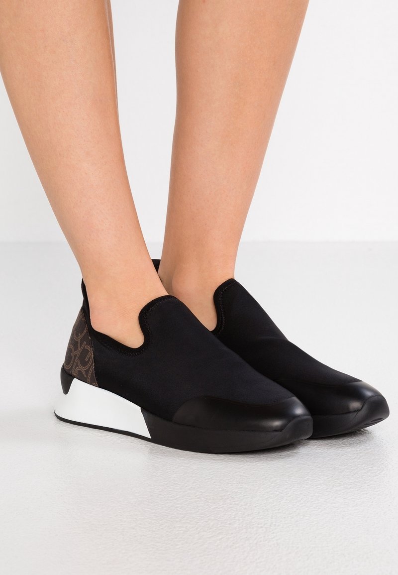 Czarne buty wsuwane z gładką, materiałową cholewką, teksturowaną brązową tylną częścią z wzorem logo oraz białą gumową podeszwą.