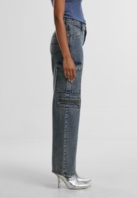 Wide-leg cargo jeans in faded blue denim, featuring side pockets and a zippered detail. Paired with metallic high-heeled shoes.