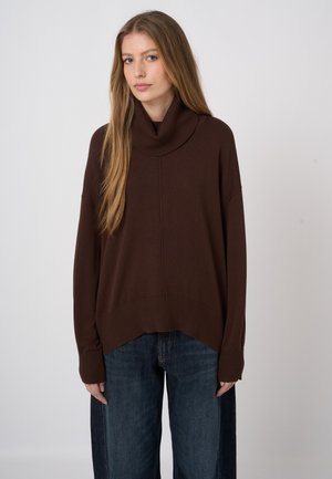 Jonge vrouw met lang blond haar die een losse donkere bruine coltrui en wijde donkerblauwe spijkerbroek draagt, staand tegen een effen achtergrond.