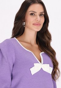 Lila Strickpullover mit V-Ausschnitt, weißem Saum und einer weißen Schleifendetail auf der Brust. Glatte Textur mit Rippmustern.
