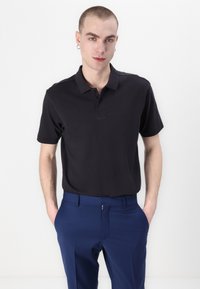 Schwarz Poloshirt mit Kragen und kurzen Ärmeln, versehen mit einem dezenten, geprägten Logo. Kombiniert mit eleganten, navyblauen Hosen.