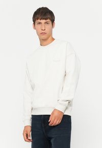 Sudadera blanca con cuello redondo y puños acanalados, que presenta un logotipo bordado tonal en el pecho. Combinada con jeans de mezclilla oscura.