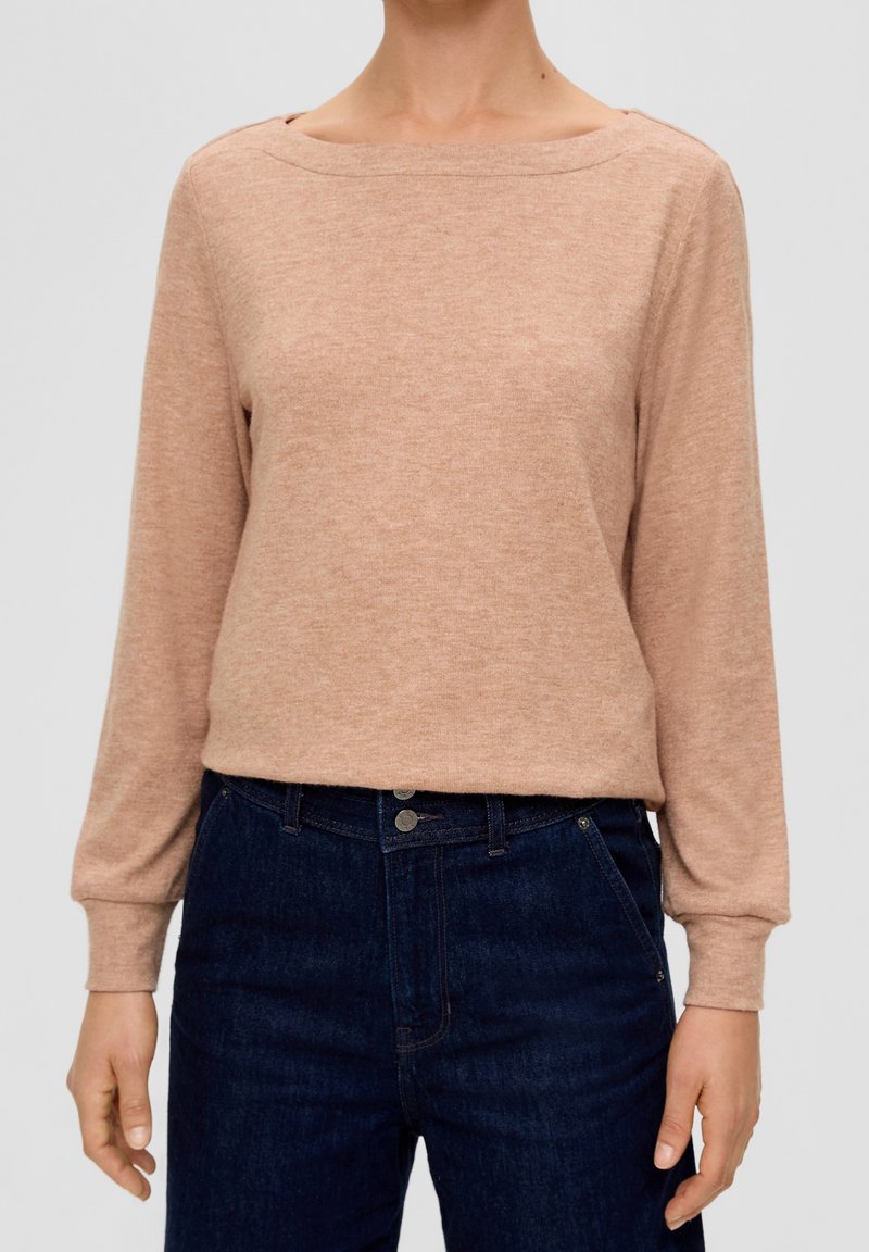 Femme portant un pull à manches longues marron clair rentré dans un jean taille haute bleu foncé, debout devant un fond uni.