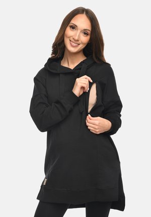 Woman in black hoodie unzipping side access opening, revealing beige underlayer, smiling with long brown hair against light background.