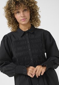 Abito nero a camicia con corpetto arricciato, chiusura frontale con bottoni e maniche a sbuffo con polsini elastici. Tessuto morbido con una forma strutturata.