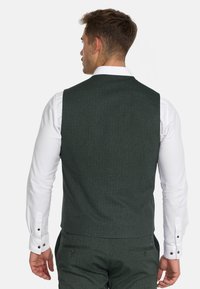 Grønn vest med fiskebensmønster og skreddersydd passform, med glatt tekstur, knappedetaljer, og en hvit langermet skjorte under.