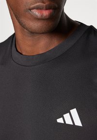 Camiseta deportiva negra con superficie texturizada y cuello redondo, que presenta un prominente logo de tres rayas blancas en el pecho.