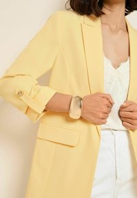 Kobieta w jasnożółtej marynarce na białej bluzce wykończonej koronką i białych spodniach, z masywną złotą bransoletą na nadgarstku.