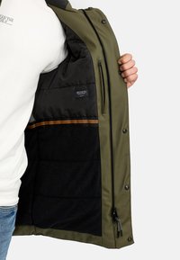 Olijfgroene jas met een gladde buitenkant, zwarte fleece voering en een horizontale bruine streep. Voorzien van ritsen en drukknoopsluitingen.