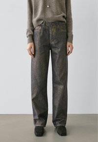 Jean en denim foncé avec une finition légèrement brillante, coupe ample, design à cinq poches et coutures contrastantes. Porté avec un pull gris et des chaussures noires.