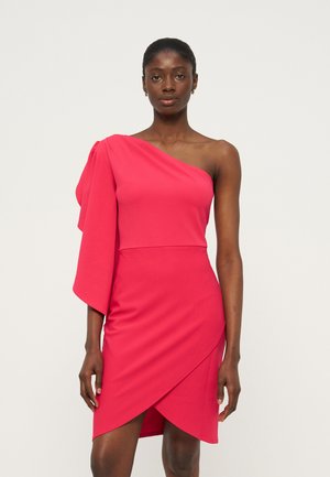 Γυναίκα που φοράει ένα φωτεινό ροζ φόρεμα με ένα ώμο, με ριγμένη μανίκι και ασύμμετρη φούστα τύπου wrapped, στέκεται μπροστά σε απλό φόντο.