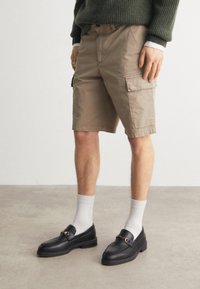 Pantaloni cargo khaki in cotone, con tasche laterali e taglio dritto. Abbinati a mocassini neri e calzini bianchi alla caviglia.