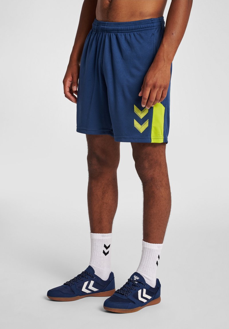 Marinblå idrottsshorts med en texturerad yta, med ett gult geometriskt mönster på sidan. Matchas med marinblå skor och vita strumpor.