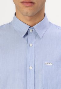Camisa de botones a rayas azul y blanca con cuello estándar, pequeño bolsillo y logo visible de "GUESS JEANS" en el bolsillo.
