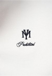 Logo "NY" entrelacé brodé en noir au-dessus du mot "Prohibited" en écriture cursive sur tissu blanc côtelé.