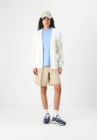 Cardigan blanc avec le numéro beige « 6 », t-shirt bleu, shorts beiges et baskets bleues. Présente une coupe décontractée avec des textures douces.