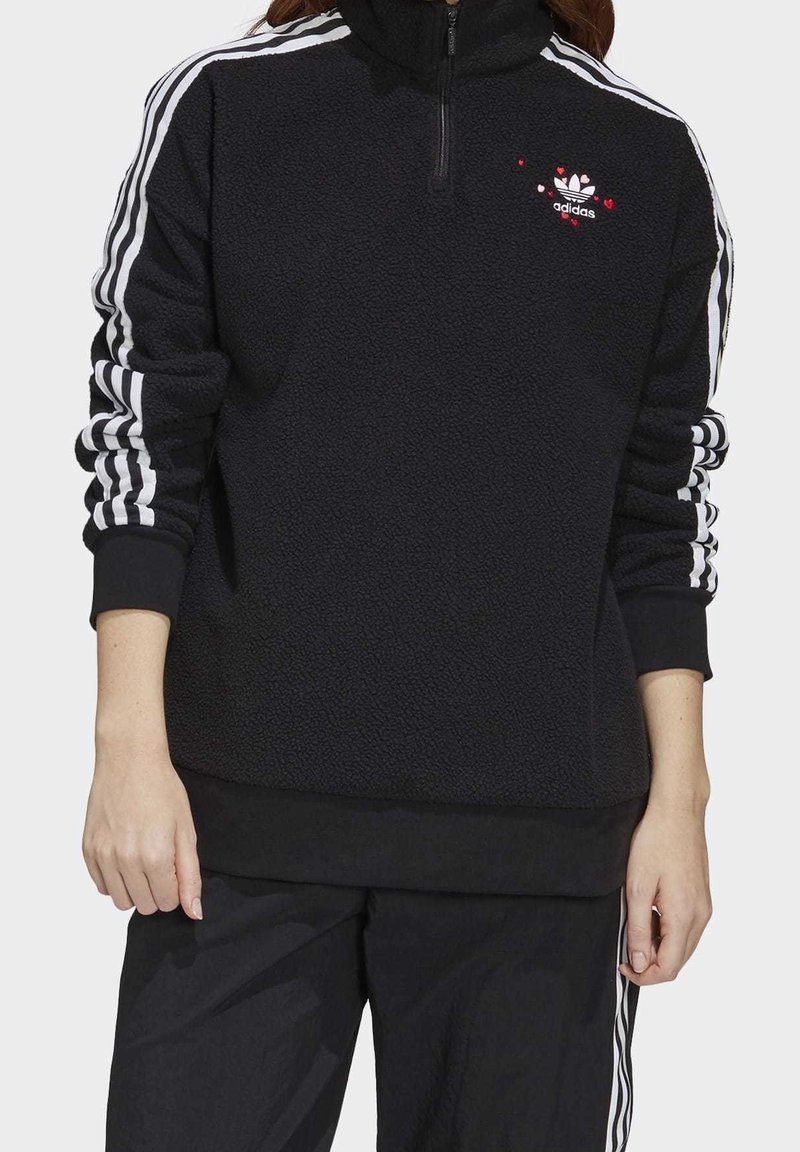 Svart fleece-pullover med hög krage, halvdragkedja och vita Adidas-ränder på ärmarna. Har broderad logotyp och hjärt-accenter.