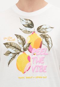 T-shirt bianco con una grafica colorata di limoni e foglie verdi, con "PEEL THE VIBE" e altro testo in caratteri audaci e giocosi.