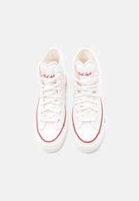 Υψηλά λευκά πάνινα παπούτσια με κόκκινες λεπτομέρειες, στρογγυλή μύτη, καουτσούκ σόλα και ραμμένα κορδόνια. Φέρουν το κεντημένο όνομα «Chuck Taylor» στη μία πλευρά.