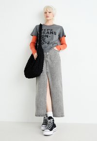Camiseta gris con texto negro, capa de manga larga naranja, falda de mezclilla gris con abertura lateral, bolsa negra texturizada, zapatillas negras altas.