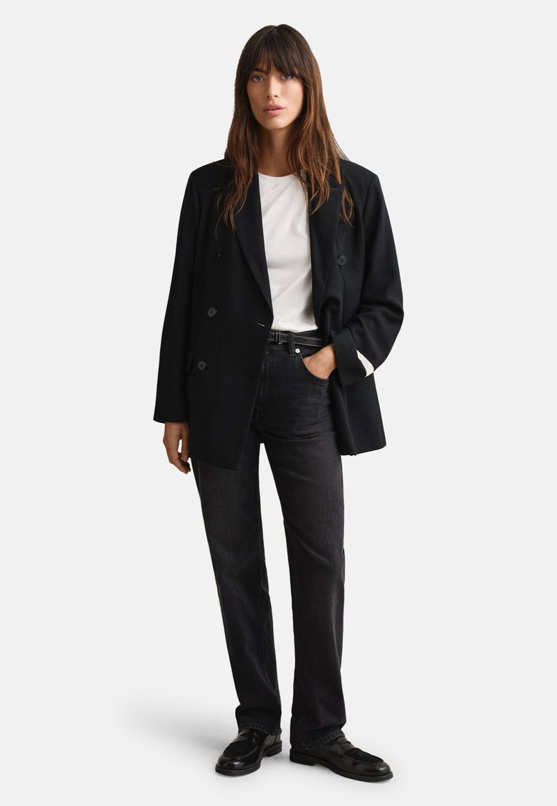 Blazer preto de duplo peito, t-shirt branca, jeans escuros, usado com sapatos pretos. Detalhes notáveis incluem botões e mangas em contraste.