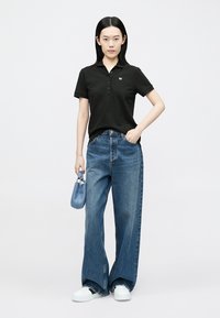 Zwart poloshirt met een kraag en drie knopen, gecombineerd met wijd uitlopende blauwe spijkerbroek en witte sneakers, met een lichtblauwe handtas in de hand.