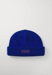 Bonnet tricoté bleu avec une texture côtelée et un rebord retourné. Présente un patch rectangulaire orange avec le logo Calvin Klein.