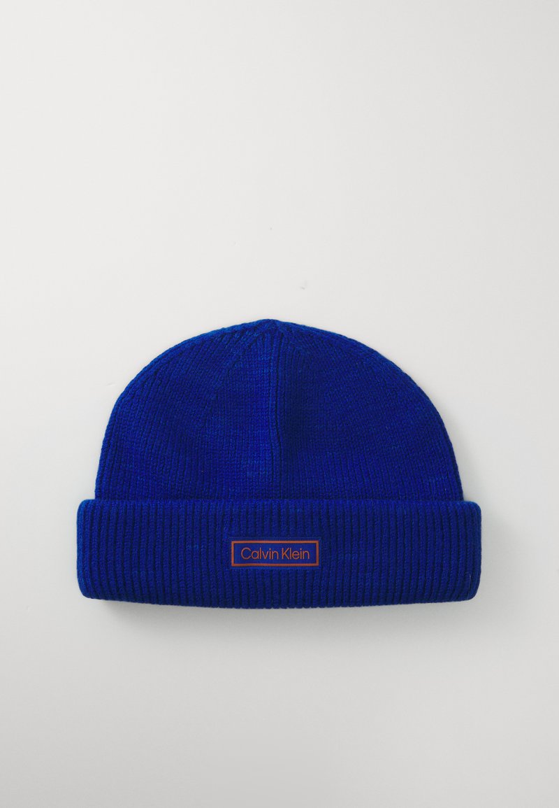 Bonnet tricoté bleu avec une texture côtelée et un rebord retourné. Présente un patch rectangulaire orange avec le logo Calvin Klein.
