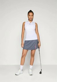 Ženska stoji v belem brezrokavnem golf majici, vzorčeni golf krilu, belih nogavicah in čevljih, ter drži golf palico pred enotno ozadjem.