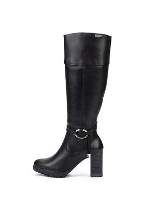 Botas negras hasta la rodilla de cuero suave. Cuentan con un tacón grueso, un acento metálico circular en el tobillo y una suela de goma plana.