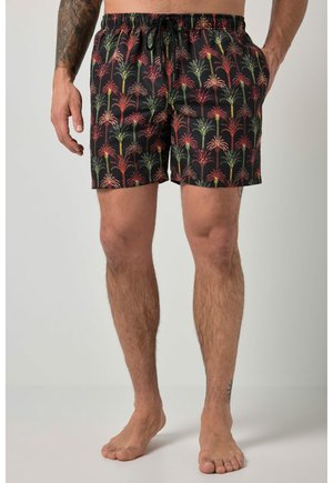 Męski tułów i nogi w czarnych kąpielówkach z czerwono-zielonym wzorem palm, stojący boso na jednolitym tle.