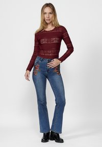Top in pizzo bordeaux a maniche lunghe abbinato a jeans in denim blu con ricami floreali sulle tasche frontali; la modella sta in piedi con sicurezza.