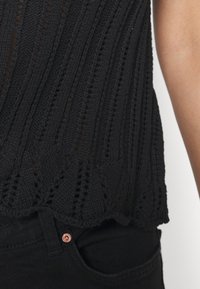 Svart stickad topp med vertikala ränder och öppet mönster. Scalloped nederkant detalj. Matchas med svarta jeans med en kopparfärgad knapp.