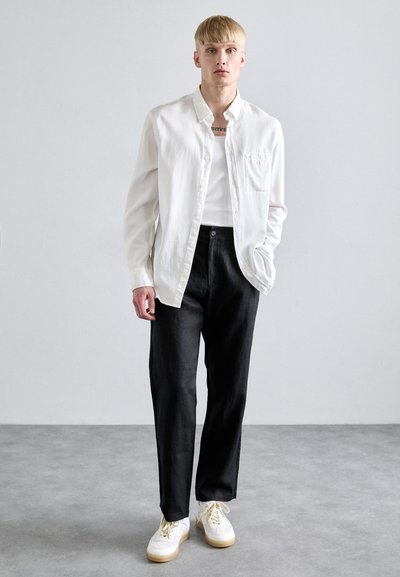 Mladý muž se světlými vlasy, oblečený v bílé košili s knoflíky, bílém tričku, černých kalhotách a bílých teniskách, stojící na šedé podlaze.