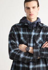 Svart og blå rutete flanellskjorte med to lommer foran, lagd over en mørkeblå hettegenser, kombinert med et svart armbåndsur.