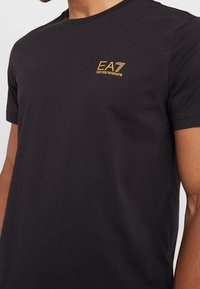 Black cotton t-shirt featuring a round neckline, short sleeves, and a gold EA7 Emporio Armani logo on the chest. Smooth texture.