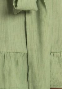 Vêtement vert clair avec une texture froissée, comportant un design plissé à l'ourlet et un détail de manche nouée.