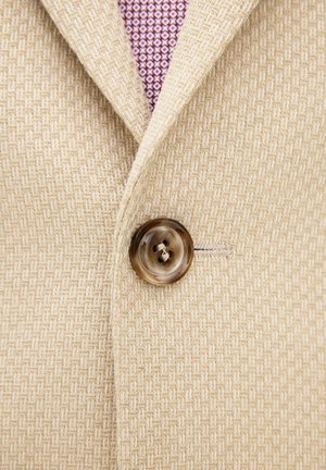 Close-up van een beige gestructureerd colbert, dichtgeknoopt over de kraag van een paars, met patroon versierd overhemd.