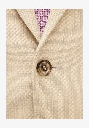 Close-up van een beige gestructureerd colbert, dichtgeknoopt over de kraag van een paars, met patroon versierd overhemd.