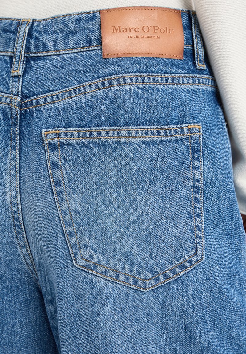 Jeans en denim bleu avec une étiquette en cuir au niveau de la taille, une poche arrière avec une couture diagonale et des surpiqûres orange contrastantes.