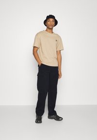 Beige Baumwoll-T-Shirt mit einem bedruckten runden Design auf der Brust, kombiniert mit marineblauen Cargo-Hosen und schwarzen Sportschuhen. Schwarzer Bucket Hat getragen.