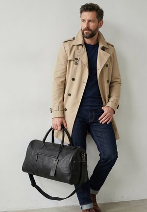 Homme en trench beige et jean foncé tenant un sac de voyage noir en cuir, debout contre un mur clair uni.